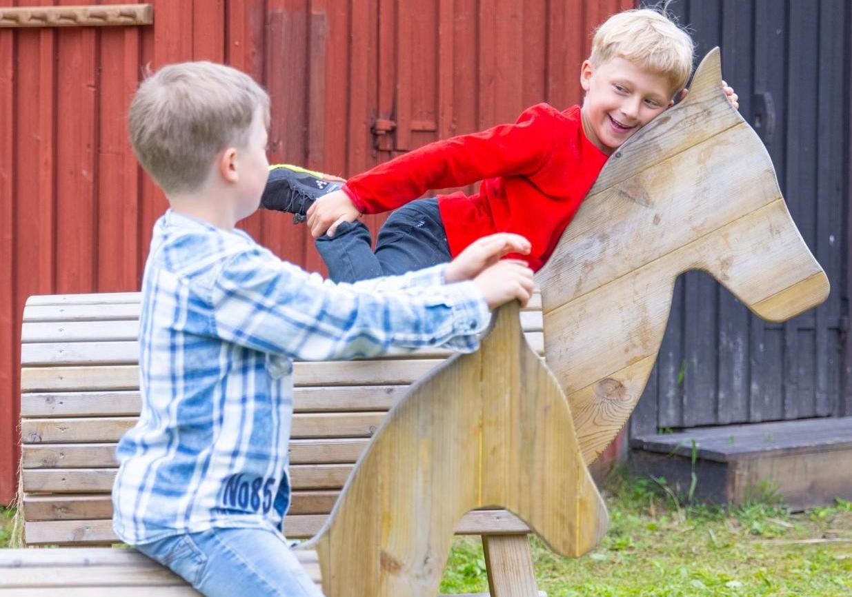 Två barn leker på trähästar utomhus. 