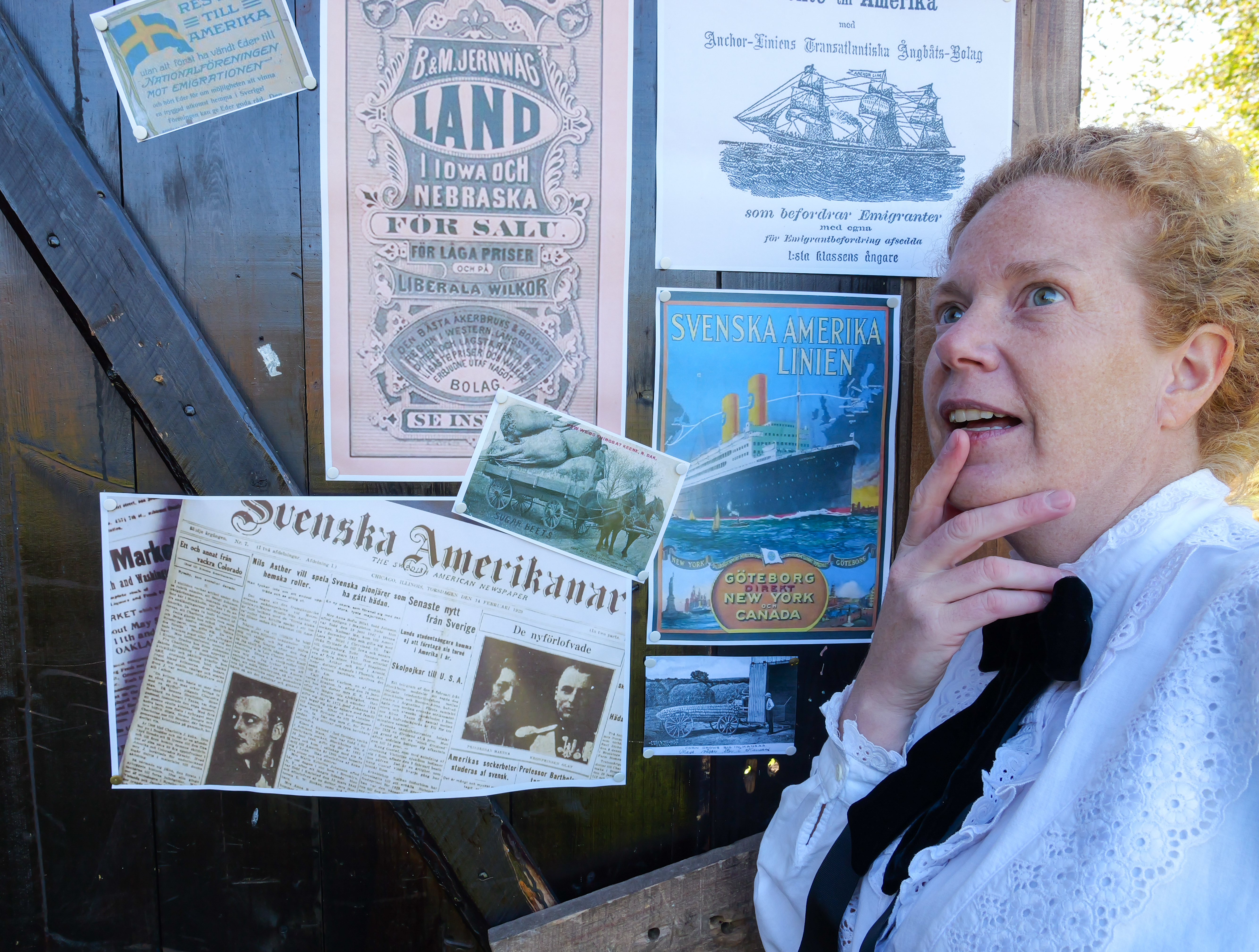 A woman dressed in old-fashioned clothes stands by some posters with information about America, looking thoughtful. 